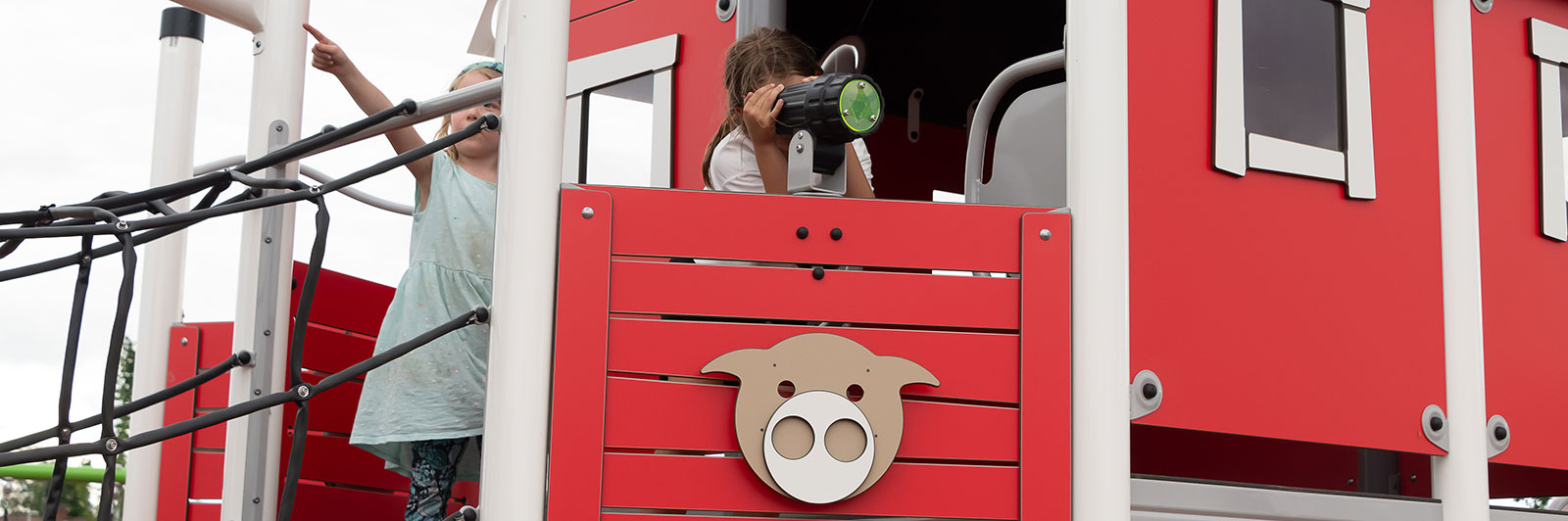 A young child interacts with a telescope on a playground unit deck. There are animal shaped panels on the side of the red playground unit.